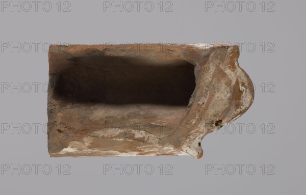 Cornice tile fragments 4, painted with a wavy twirl pattern; tile cullet; stoves; plant decorations; 19th century; Hutsul region, Poland., A weathered, hollow ceramic or terracotta object shown from above with a rectangular cavity and an ornate rounded end, displaying rough, uneven surfaces and chips. The visible colors include various shades of brown and reddish-brown, patches of white and off-white, small areas of dark brown or black inside the cavity, and tiny spots of green near the ornate edge. The background is a uniform light gray.
