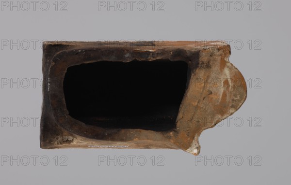 Cornice tile fragments, four; maker, tile cullet; stoves; plant decorations; Hutsul region; painted with a wavy twirl pattern; 19th century; Poland., A rectangular hollow object with weathered, rough surfaces and an irregular oval opening occupies the center of the image, showing an interior that appears very dark. The exterior surfaces show mottled shades of brown, ochre, tan, and gray with patches of lighter beige and occasional darker brown areas, and there is a rounded protrusion on the right side with similar coloring. The background is a uniform pale gray.