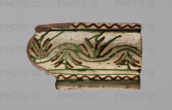 Cornice tile fragments 4, painted with a wavy twirl pattern; tile cullet; stoves; plant decorations; 19th century; Hutsul region, Poland., A fragment of a ceramic shard with a curved profile resting against a neutral gray background, showing a glazed surface in off-white with visible crackle and areas of exposed brown clay at the edges; the decoration consists of flowing, organic brushstrokes in green and brown forming wave-like and leaf-like motifs across the shard, with a wavy brown border near the top and bottom edges and a darker green line accenting a raised ridge; the surface shows spots of discoloration and wear.