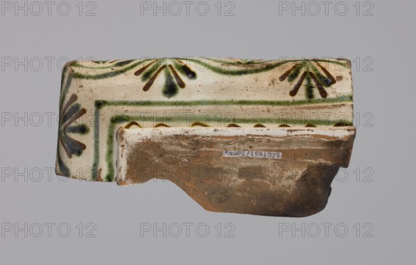 Cornice tile fragments 4, tile cullet; painted with a wavy twirl pattern; 19th century; Hutsul region, Poland; plant decorations., Function: cornice tile., A broken ceramic fragment with a glazed upper surface showing a cream background and decorative brushstrokes in green and dark brown, with some areas of blue-green; the lower exposed clay is brown and unglazed with some white residue; the fragment has a curved profile and worn edges, and the overall image background is a flat light gray.