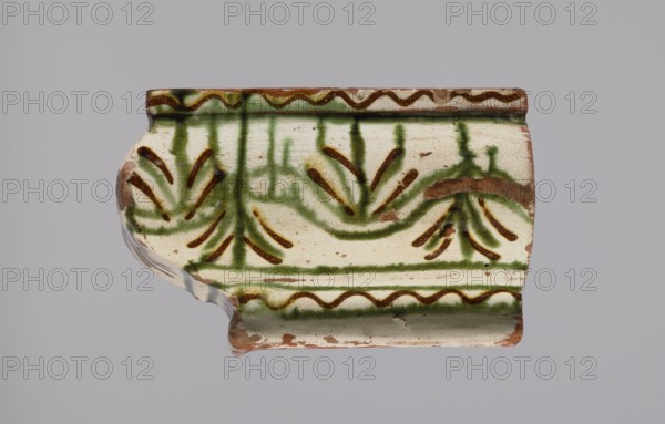 Cornice tile fragments 4, painted with a wavy twirl pattern; tile cullet; stoves; plant decorations; 19th century; Hutsul region, Poland., A fragment of a ceramic object rests against a plain light gray background, showing an off-white surface with green and brown painted decorative motifs; the piece has a curved profile with a raised rim along the top and bottom edges, the top rim featuring a wavy brown line bordered by green, and the body decorated with green vertical strokes and curved green bands accented by brown dash-like marks that form floral or fan-like shapes; there are areas of exposed reddish-brown clay where the glaze is chipped, and the glaze surface shows fine crackling and small dark specks.