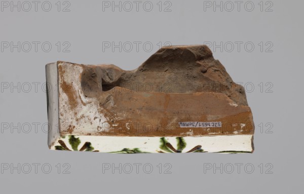 Cornice tile — fragments, four;, tile cullet; stoves; plant decorations; Hutsul region; painted with a wavy twirl pattern; 19th century; Poland., A broken piece of pottery or ceramic with an irregular top surface in shades of brown and tan, a lower band of white with green and dark brown decorative brushstrokes, and some areas of off-white and gray; the object rests against a plain gray background.