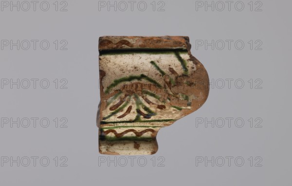 Cornice tile — fragments, four; maker, tile cullet; stoves; plant decorations; Hutsul region; tiles; painted with a wavy twirl pattern; 19th century; Hutsul region, Poland; tile cullet, A fragment of a ceramic object with a worn, irregular shape shows a surface of beige and light tan with areas of exposed reddish-brown clay; decorative brushstrokes in green and dark brown form curved and linear patterns across the face, and there are traces of darker green along raised ridges; the piece has a rounded protruding section on one side and a flat stepped edge on the opposite side, with patches of dirt and small chips visible on the surface against a pale gray background.