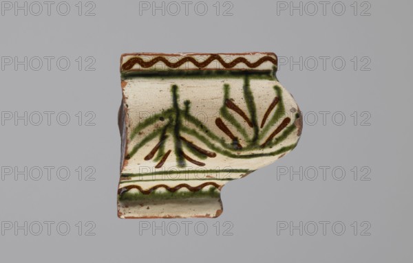 Cornice tile fragments 4, tile cullet; painted with a wavy twirl pattern; 19th century; Hutsul region, Poland; stoves; plant decorations., A fragment of a ceramic tile with a cream-colored glazed surface showing a decorative pattern in green and brown; the shard has a curved edge and a molded profile with a raised band at the top and bottom, each band edged by a zigzag brown line and green shading, and the central field displays stylized plant-like motifs rendered in green strokes with brown accents; the ceramic body visible where glaze is chipped appears reddish-brown.