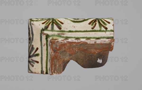 Cornice tile fragments 4, tile cullet; painted with a wavy twirl pattern; 19th century; Hutsul region, Poland; stoves, plant decorations., A broken ceramic fragment with glazed decoration sits against a plain gray background, showing a creamy white surface with green painted lines and stylized radial motifs in green and brown, and an exposed unglazed terracotta interior that is reddish-brown with patches of darker brown and worn areas, plus a small patch of white residue on the unglazed section.