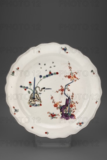 Platter with a yellow lion, Koenigliche Porzellain Fabrique, Meissen ca. 1740, kakiemon style, mit gelbem Loewe pattern. Platter with a wavy edge collar, slightly convex profiled roller, brown rim; in the mirror, Kakiemon-type decoration: a tiger creeping around a bamboo shoot, a blossoming plum bush and small sprays of flowers and leaves, some with blemishes; tiger with a golden eye. Porcelain dish, height 6 cm, diameter 38 cm; Saxony, Meissen., A white scalloped ceramic plate with a thin dark rim sits upright on a transparent stand against a gray background, decorated with a group of colorful painted motifs including a purple sculptural form at one side beside a blue branch with red clustered blossoms, a nearby arrangement with a vertical blue element and a pale yellow striped creature or ornamental knot, and scattered small floral sprays and dots in red, blue, green, and teal across the interior and along the rim.