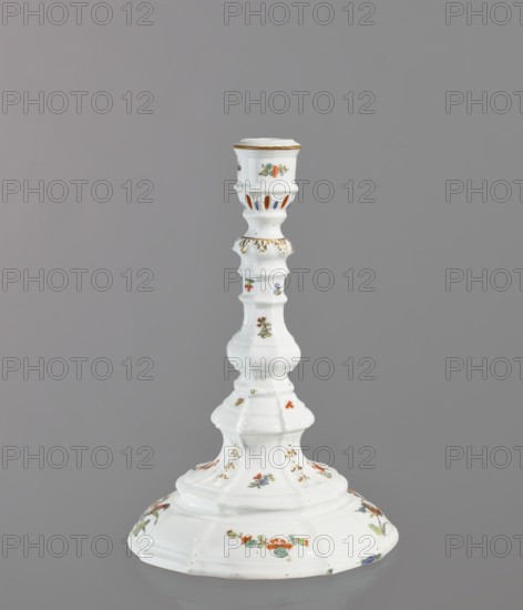Candlestick, Koenigliche Porzellain Fabrique, Meissen 1710–1763. One-candle candlestick with profiled baluster shaft and 8-segment round foot; candle socket mounted on a profile with relief acanthus leaves. Decorated with gilding acanthus frieze, relief campanulas and twigs and projections of Far Eastern flowers; two twigs with a bird around the foot; two small insects on the socket. Brown border around the edge of the candle holder. Circa 1750. Meissen, Saxony. Function: lighting. Height 23.8 cm; diameter 16 cm., A white ceramic candlestick with a glossy finish standing against a plain gray background, featuring a flared base with a scalloped edge and a tall, tiered stem with rounded and angular segments; the surface is decorated with small floral motifs in green, orange, blue, and red, along with gold accents on raised details and the top rim, and the overall shape shows a gently curving silhouette with decorative cutouts near the top.