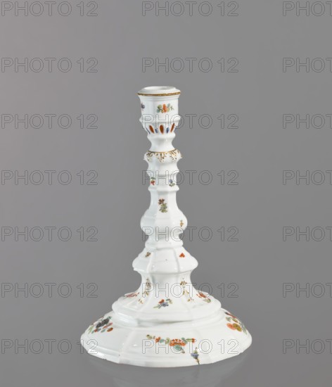 Candlestick, Königliche Porzellan-Manufaktur Meissen ca. 1750. One-candle candlestick with profiled baluster shaft and 8-segment round foot; candle socket mounted on a profile with relief acanthus leaves. Decorated with gilding acanthus frieze, relief campanulas, twigs and projections of Far Eastern flowers, two twigs with a bird around the foot, and two small insects on the socket; brown border around the edge of the candle holder. Porcelain, Meissen, Saxony; height 23.8 cm, diameter 16 cm., A white ceramic candlestick with a glossy finish standing against a plain gray background, featuring a flared base with a gently scalloped edge decorated with small floral motifs in orange, green, blue, and yellow, a turned column with a series of rounded and ridged sections also adorned with tiny floral decorations in similar colors and touches of gold trim, a midsection with delicate gold-painted accents, and a cup-shaped top with vertical cutouts and additional floral motifs and gold edging.