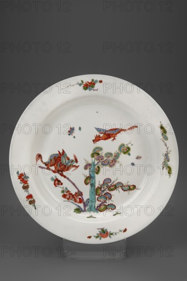 Platter with flying dragons, Koenigliche Porzellain Fabrique, Meissen ca. 1730, Kakiemon style; round platter with smooth collar and Kakiemon-type decoration: in the mirror two flying, attacking dragons — one above a blooming bamboo shoot, the other sitting on a withered bamboo shoot with a peony flower next to it; on the collar four branches of Indian flowers. Porcelain vessel, height 2.5 cm, diameter 38 cm. Inventory no. 124297; war loss vase with lid., A round white ceramic plate with a wide rim and shallow well decorated with painted floral and bird motifs in colors including red, green, blue, purple, turquoise, and gold tones; the central scene shows a stylized tree or plant with a turquoise trunk and multicolored swirling foliage, accompanied by two bird-like creatures in red and blue perched and flying above, while the rim features several small floral sprays and decorative motifs in red, green, and blue spaced around the edge; the plate sits on a clear display stand against a d...