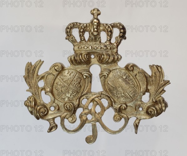 Lock plate, ca. 1750. Brass furniture fitting, height 9 cm, width 9.8 cm, thickness 1 cm. Decoration: crowns, Saxony electoral/Wettyn coat of arms, coat of arms of the Polish-Lithuanian Commonwealth, plant decoration., A decorative metallic ornament with a gold color finish, shaped with a central crown at the top and ornate scrolling elements below; the crown has rounded arches with bead-like details and rests above a pair of rounded medallion shapes with textured, embossed surfaces. On either side of the medallions are symmetrical wing-like leaf flourishes and curled scrollwork, creating a balanced, baroque-style composition. The piece shows subtle variations in gold tone and a slightly reflective surface against a pale, neutral background that appears off-white to light gray.
