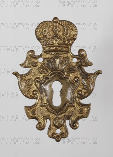 Lock plate, maker, attributed to Léopold Méyet 1850–1912. Crowns and plant decoration; furniture fitting. Late 17th–early 18th century, Central Europe possibly Saxony. Height 8.6 cm, width 6.2 cm, depth 0.25 cm., A decorative ornate metal escutcheon with an elaborate symmetrical shape and a central keyhole, showing a crown-like top and scrolling floral and leaf motifs on its sides and bottom; the surface appears metallic with a gold color and darker patina in recessed areas, set against a plain light gray background.
