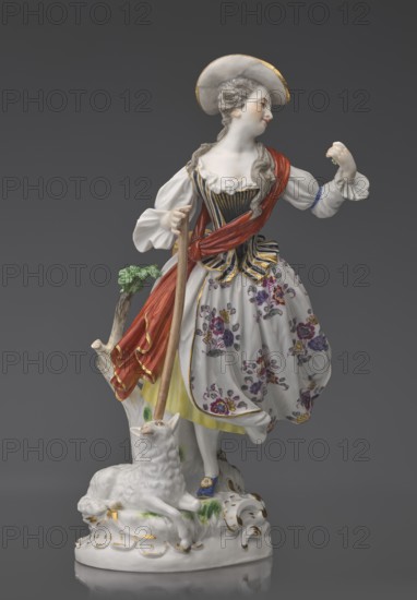 Shepherdess with a Sheep, Kaendler, Johann Joachim 1706–1775, sculptor; Koenigliche Porzellain Fabrique, Meissen 1710–1763, manufacturer. Porcelain figurine on an oval base with rocaille and a tree trunk with leafy branch: a young woman in a dancing running? pose wearing a flowing flowered half-length skirt, black-and-gold striped bodice with a small handbag tied at the back below a basque, blouse with puffed sleeves tied at the elbow with a blue ribbon, and a red sash with long ends loosely thrown over the left shoulder and tied at the waist. Medium-length pinned-up hair falling in curls down her back, neckline, and shoulder; she wears a hat with a blue ribbon. She holds a shepherd’s staff upper part whipped resting on the head of a sheep lying at her feet; her head is tilted slightly left and she holds a flower whipped in her raised left hand. 19th/20th century after a model ca. 1750. Saxony, Meissen. Height 23.5 cm., A porcelain figure of a woman wearing a wide-brimmed hat with gold...