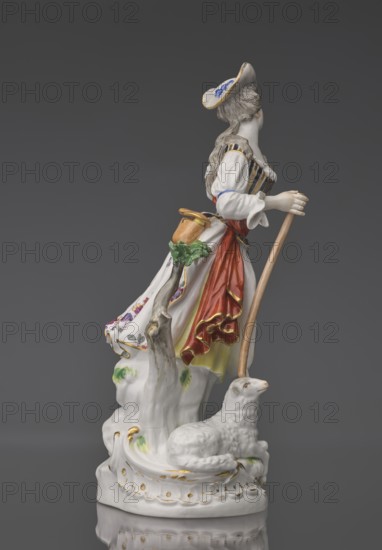 Shepherdess with a Sheep, Kaendler, Johann Joachim 1706–1775, sculptor; Koenigliche Porzellain Fabrique, Meissen 1710–1763, manufacturer. Porcelain figurine on an oval base with rocaille and a tree trunk with a leafy branch: a young woman in a dancing running? pose wearing a flowing flowered half-length skirt, black-and-gold striped bodice, small handbag tied at the back below a basque, blouse with puffed sleeves tied at the elbow with a blue ribbon; a red sash with long ends loosely thrown over the left shoulder and tied at the waist. Medium-length pinned-up hair falling in curls; she wears a hat with a blue ribbon. She holds a shepherd's staff upper part whipped resting on the head of a sheep lying at her feet. Her head is tilted slightly left and she holds a flower whipped in her raised left hand. 19th/20th century after a model ca. 1750, Saxony, Meissen. Figurine, height 23.5 cm., A porcelain figurine of a woman standing on a decorative base next to a reclining sheep, with the woma...