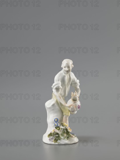 Gardener, Koenigliche Porzellain Fabrique, Meissen 1710–1763; Kaendler, Johann Joachim 1706–1775 sculptor? ; Reinicke, Peter 1715–1768 sculptor?; gardeners; figurine. On a circular base with a high tree trunk and plastic tufts of flowers stands a slightly bent barefoot man, leaning against the trunk, in knee-length trousers and a luxurious shirt with rolled-up sleeves; he holds a watering can in his left hand, tilting it to water the flowers, and supports the spout with a strainer in the palm of his other hand. ca. 1760. Meissen, Saxony, Germany. Porcelain figurine; height 12 cm., A small porcelain figure of a person with pale skin and light hair stands on a white base decorated with green leaves and flowers in blue, yellow, pink, and white; the figure wears a white top with rolled sleeves and a pale yellow skirt, holds a white jug with floral decoration and a gold rim, and leans against a white draped support, all set against a neutral gray background.