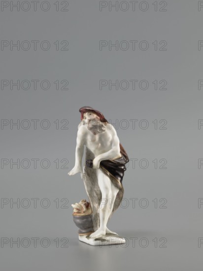 Winter — Allegory. Meissen porcelain Koenigliche Porzellain Fabrique, 1710–1763; mid-18th century, Saxony, Meissen. Figurine, black on a flat quadrangular pedestal; height 11.5 cm. Depicts an old naked, barefoot bearded man standing in contrapposto, covered with a fur-lined cloak, a horn thrown over his head and held in his left hand, leaning slightly and warming his right hand over a cauldron with a flame at his feet. Genres/subjects: seasons allegory, winter; figurine., A small ceramic figurine of a standing bearded man wearing a hat and a draped cloth, with pale white skin tones, brown hat and cloth with darker brown and black shading, grayish and beige accents on a textured vertical element behind him, and a grayish vessel with beige and brown contents at his feet, all set against a plain light gray background.