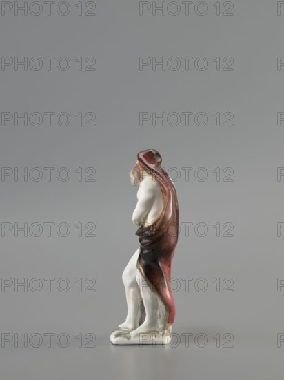 Winter — Allegory. Meissen porcelain Koenigliche Porzellain Fabrique, Meissen, ca. 1710–1763. Seasons allegory: Winter. Figurine, mid-18th century, Meissen, Saxony. Black glaze on a flat quadrangular pedestal. Depicts an old naked, barefoot bearded man standing in contrapposto, covered with a fur-lined cloak, a horn thrown over his head and down his back and held in his left hand; he leans slightly and warms his right hand over a cauldron with a flame at his feet. Height 11.5 cm., A small ceramic figure on a plain surface against a gray background, showing a standing human form draped in a long cloak. The figure is mostly white with flesh-toned areas for the exposed limbs and head, and the cloak displays shades of deep red, brown, and darker maroon with some blackish shading; the base is white with a touch of grayish discoloration. The figure's posture is slightly hunched with arms drawn in and the cloak falling to the ground, and the surface has a glossy finish.