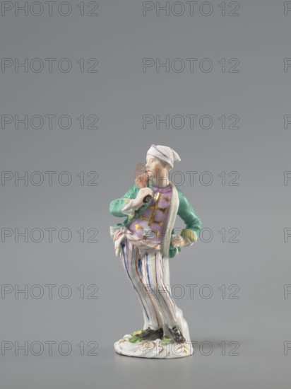 Man with a Potato Masher, Reinicke, Peter 1715–1768 modeler?, Koenigliche Porzellain Fabrique, Meissen 1710–1763 manufacturer. On a flat circular pedestal with rocaille merging into a tree trunk stands a young man leaning on the trunk at waist level. In contrapposto, with his head turned to the right, he holds a short oval board with a handle—an instrument for stewing potatoes—resting on his right shoulder. Dressed in trousers with colorful thin stripes tied loosely at the waist with a belt, black shoes with a buckle, a short green jacket with box pleats, a pink waistcoat with gold braiding, and a soft cap similar to a nightcap. Ca. 1755, Saxony; Meissen. Figurine, height 13.5 cm., A small porcelain figure of a person standing on an ornate base, wearing a white headscarf with a thin colored band, a green jacket with yellow cuffs over a purple vest, and white striped trousers with vertical colored lines; the figure holds a brown object near the face with one hand while the other rests o...
