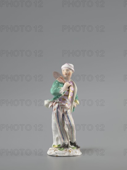 Man with a Potato Masher, Reinicke, Peter 1715–1768 modeler?, Koenigliche Porzellain Fabrique, Meissen 1710–1763 manufacturer, figurine. On a flat, circular pedestal with rocaille merging into a tree trunk, stands a young man leaning on the trunk at waist level. In contrapposto, head turned to the right, he holds a short oval board with a handle—an instrument for stewing potatoes—resting on his right shoulder. Dressed in trousers with colorful thin stripes tied loosely at the waist with a belt, loose ends, black shoes with a buckle, a short green jacket with box pleats, a pink waistcoat, with gold braiding and a soft cap similar to a nightcap. Ca. 1755, Saxony; Meissen; figurine; height 13.5 cm., A small painted porcelain figurine of a standing person wearing a white cap with pink trim, a green jacket, a pink vest, and white trousers with vertical blue and pink stripes, holding a brown object over one shoulder and leaning against a white tree stump, mounted on a white decorative base w...