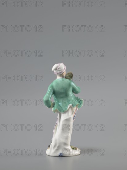 Man with a Potato Masher, Reinicke, Peter 1715–1768 modeler?; Koenigliche Porzellain Fabrique, Meissen 1710–1763 manufacturer. On a flat, circular pedestal with rocaille merging into a tree trunk stands a young man leaning on the trunk at waist level. In contrapposto, head turned to the right, he holds a short oval board with a handle—an instrument for stewing potatoes—resting on his right shoulder. Dressed in trousers with colorful thin stripes tied loosely at the waist with a belt, black shoes with a buckle, a short green jacket with box pleats, a pink waistcoat with gold braiding, and a soft cap similar to a nightcap. Ca. 1755, Saxony; Meissen. Figurine; height 13.5 cm., A small porcelain figurine shown from the back on a gray background, featuring a figure wearing a white head covering with thin pink stripes, a green jacket with a flared hem, and a long white skirt with subtle colored stripes at the side; the figure holds a small brownish object near the shoulder and stands on a wh...
