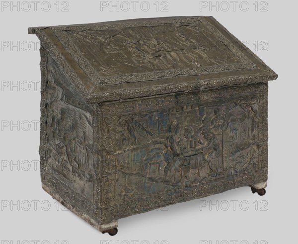 Box with straight walls and a diagonally sloping lid, covered with embossed brass sheet depicting feast scenes in an inn; mounted on four steel rollers. Walls, bottom and lid made of boards; rollers attached to two wide strips glued to the bottom on the short sides. 2nd half of the 19th century. Central Europe, possibly Germany Silesia. Historicist style; subject: inns, feasts. Piece of furniture storage. Height 49 cm, width 60.5 cm, depth 32.7 cm., and origin; inscriptions/author unknown., The image shows a rectangular metal chest with a sloped hinged lid, covered in raised decorative relief scenes and ornate border patterns; the surface appears mostly dark bronze with areas of greenish and bluish patina and hints of brown, and the chest rests on small rounded feet with a slightly lighter base edge.