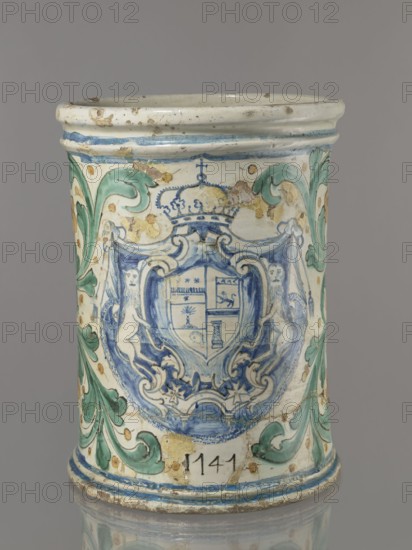 Vessel - flowerpot; cache-pot vessel; cylindrical flowerpot with profiled rollers at base and spout. Decorated throughout with a stylized fleshy plant twig in the form of an acanthus with flowers. On the front is a cartouche with a five-field coat of arms beneath a royal crown; the elaborate cartouche is supported by two lions rampant, with a griffin holding a pennant. Below the coat of arms is the date 1747. Green plant decoration with ochre flowers; blue cartouche shades from navy to light blue. A hole in the bottom for draining water. 1747. Italy, Caltagirone . Height 74 cm; width 31 cm., A cylindrical ceramic vessel with a glossy finish displaying decorative painted motifs in blue, green, white, and beige; the top rim is off-white with small brown spots and a thin blue band below it, and the body features a large central blue ornamental shield flanked by stylized leaf scrolls in green and beige, with traces of wear and chipped glazed areas exposing underlying beige clay, and a few ...