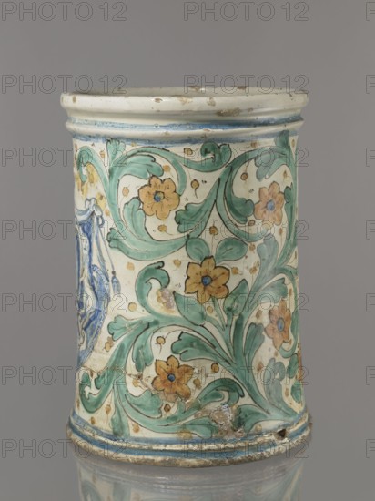 Vessel - flowerpot, unspecified manufacture; coats of arms; cartouches; cache-pot vessel; plant decorations. Cylindrical flowerpot with profiled rollers at the base and spout. Decorated throughout with a motif of a stylized, fleshy plant twig in the form of an acanthus with flowers. On the forehead is a cartouche with a five-field coat of arms beneath a royal crown. The elaborate cartouche is supported by two lions rampant, with a griffin holding a pennant. Below the coat of arms is the date 1747. Green plant decoration with ochre flowers; blue cartouche shades ranging from navy blue to light blue. A hole in the bottom for draining water. 1747; Italy, Caltagirone . Vessel, height 74 cm, width 31 cm., A cylindrical ceramic vessel with a pale background decorated with a flowing green leafy pattern and a group of yellow-orange flowers with blue centers, bordered at top and bottom by thin blue bands, showing areas of surface wear and small chips, against a neutral gray backdrop.