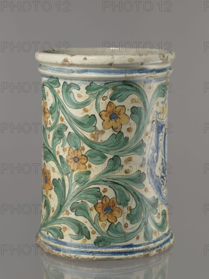 Vessel - flowerpot, cache-pot vessel. Cylindrical flowerpot with profiled rollers at the base and spout. Decorated throughout with a stylized, fleshy plant twig motif in the form of an acanthus with flowers. On the front is a cartouche with a five-field coat of arms beneath a royal crown; the elaborate cartouche is supported by two lions rampant, with symbols of a griffin holding a pennant. Below the coat of arms is the date 1747. Painted green plant decoration with ochre flowers; blue cartouche shades ranging from navy blue to light blue. A hole in the bottom for draining water. 1747. Italy, possibly Caltagirone. Height 74 cm, width 31 cm., A cylindrical ceramic vessel with a glossy cream background showing decorative green scrolling leaves and mustard-yellow flowers with blue centers, framed by thin blue bands near the top and bottom; the piece shows areas of glaze wear and small chips, and the color palette includes cream, green, mustard-yellow, blue, and touches of brown.