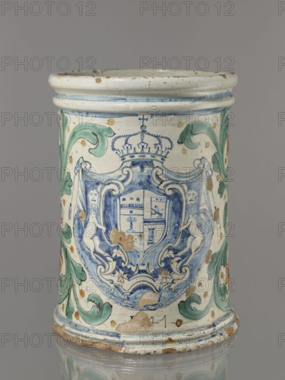 Vessel - cache-pot flowerpot in the shape of a cylindrical flowerpot with profiled rollers at the base and spout. Decorated throughout with a stylized, fleshy plant twig in the form of an acanthus with flowers. On the forehead is a cartouche with a five-field coat of arms beneath a royal crown. The elaborate cartouche is supported by two lions rampant, with a griffin holding a pennant. Below the coat of arms is the date 1747. Green plant decoration with ochre flowers, blue cartouche shades ranging from navy blue to light blue. A hole in the bottom for draining water. 1747. Italy, possibly Caltagirone. Height 74 cm, width 31 cm., A cylindrical ceramic object with a glossy off-white surface decorated with blue and green painted motifs, showing a central blue coat of arms flanked by two stylized creatures and topped with a crown, surrounded by flowing green acanthus-like leaves and scattered brownish spots and worn areas revealing a darker clay underneath, with a rounded rim and a molded ...