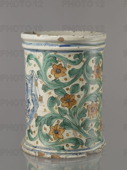 Vessel - flowerpot, cache-pot vessel. Vessel in the shape of a cylindrical flowerpot with profiled rollers at the base and spout. Decorated throughout with a stylized, fleshy plant twig in the form of an acanthus with flowers. On the forehead is a cartouche with the five-field coat of arms beneath a royal crown. The elaborate cartouche is supported by two lions rampant, with symbols of a griffin holding a pennant. Below the coat of arms is the date 1747. Green plant decoration with ochre flowers, blue cartouche shades ranging from navy blue to light blue. A hole in the bottom for draining water. 1747, Italy, Caltagirone, height 74 cm, width 31 cm. Manufacturer unspecified., A cylindrical ceramic vessel with a rounded rim and base, showing areas of wear and small chips, decorated with a scrolling floral pattern; the decoration includes green leafy scrolls and orange flowers with blue centers set against a cream background, with additional blue painted bands near the rim and base and sma...