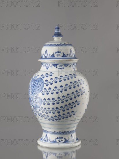 Vase with lid, Fayence-Manufaktur Muenden faience factory; 1732–1854, architecture, Méyet, Leopold 1850–1912; birds, plant motifs. Baluster vase with a short, slightly flared neck, pear-shaped belly, on a conical foot, with a domed lid terminating in a conical handle. Around the lid, neck, and foot is a pattern of triple semicircles, fir-tree and oval plant motifs and borders. On the front, in a circular cartouche, an architectural view with two towers and three houses with gable roofs. Cartouche topped with a plant palmette with a circular motif at the bottom and richly ornamented on both sides with a scale mosaic, Chinese latticework, double volutes, and two birds at the top. Branches with flowers-fruits or oak leaves radiate horizontally from the cartouche. On the back are small stars and a bipartite shoot motif. 3rd quarter of the 18th century, Germany; Hannoversch Münden. Vessel; height 41.8 cm, width 21.0 cm., A porcelain lidded jar with a rounded body and a tapered base displaye...