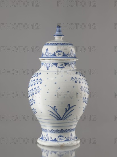 Vase with lid, Fayence-Manufaktur Muenden faience factory; 1732–1854, Hannoversch Münden; maker: Méyet, Leopold 1850–1912. Baluster vase with a short, slightly flared neck, pear-shaped belly, on a conical foot, with a domed lid terminating in a conical handle. Around the lid, neck, and foot is a pattern of triple semicircles, fir-tree and oval plant motifs and borders. On the front, in a circular cartouche, an architectural view with two towers and three houses with gable roofs; the cartouche is topped with a plant palmette with a circular motif at the bottom and richly ornamented on both sides with a scale mosaic, Chinese latticework, double volutes, and two birds at the top. Branches with flower-fruits or oak leaves radiate horizontally from the cartouche. On the back are small stars and a bipartite shoot motif. Birds and plant motifs. 3rd quarter of the 18th century, Germany; Hannoversch Münden. Vessel. Height 41.8 cm; width 21.0 cm., A lidded ceramic jar with a rounded body and a f...