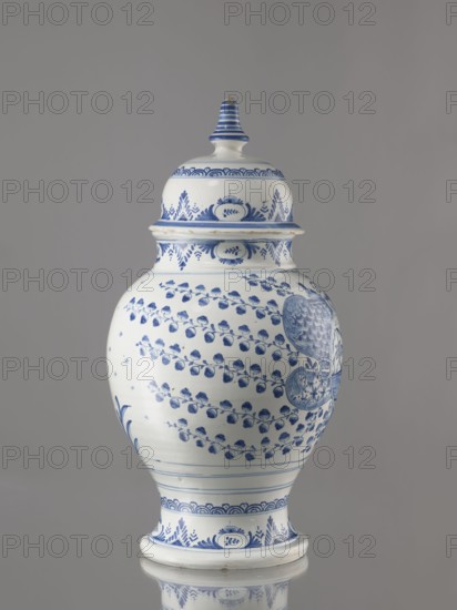 Vase with lid, Fayence-Manufaktur Muenden faience factory; 1732–1854, architecture, Méyet, Leopold 1850–1912. Baluster vase with a short, slightly flared neck, pear-shaped belly, on a conical foot, with a domed lid terminating in a conical handle. Around the lid, neck, and foot is a pattern of triple semicircles, fir-tree and oval plant motifs and borders. On the front, in a circular cartouche, an architectural view with two towers and three houses with gable roofs. The cartouche is topped with a plant palmette with a circular motif at the bottom and richly ornamented on both sides with a scale mosaic, Chinese latticework, double volutes, and two birds at the top. Branches with flowers, fruits, or oak leaves radiate horizontally from the cartouche. On the back are small stars and a bipartite shoot motif. 3rd quarter of the 18th century, Hannoversch Münden, Germany. Vessel; height 41.8 cm, width 21.0 cm., A lidded ceramic jar with a rounded body and flared base sits against a neutral gr...
