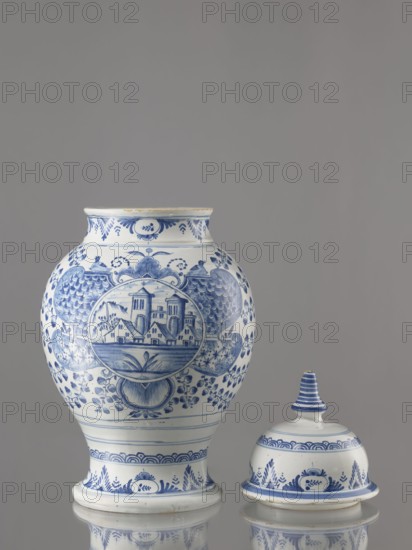 Vase with lid, Fayence-Manufaktur Muenden faience factory; 1732–1854 Hannoversch-Münden, architecture, Méyet, Leopold 1850–1912; birds, plant motifs. Baluster vase with a short, slightly flared neck, pear-shaped belly, on a conical foot, with a domed lid terminating in a conical handle. Around the lid, neck, and foot is a pattern of triple semicircles, fir-tree and oval plant motifs and borders. On the front, in a circular cartouche, an architectural view with two towers and three houses with gable roofs. The cartouche is topped with a plant palmette with a circular motif at the bottom and richly ornamented on both sides with a scale mosaic, Chinese latticework, double volutes, and two birds at the top. Branches with flowers–fruits or oak leaves radiate horizontally from the cartouche. On the back are small stars and a bipartite shoot motif. 3rd quarter of the 18th century, Germany; Hannoversch-Münden. Vessel; height 41.8 cm, width 21.0 cm., A ceramic jar and its lid sit against a plai...