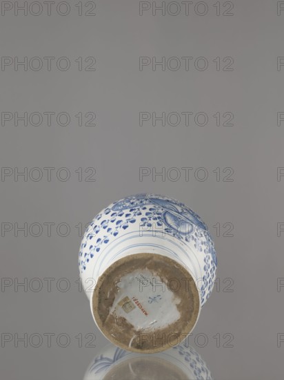 Vase with lid, Fayence-Manufaktur Muenden faience factory; 1732–1854 Hannoversch-Münden, Méyet, Leopold 1850–1912. Baluster vase with a short, slightly flared neck, pear-shaped belly, on a conical foot, with a domed lid terminating in a conical handle. Around the lid, neck, and foot is a pattern of triple semicircles, fir-tree and oval plant motifs and borders. On the front, in a circular cartouche, an architectural view with two towers and three houses with gable roofs; the cartouche is topped with a plant palmette with a circular motif at the bottom and richly ornamented on both sides with a scale mosaic, Chinese latticework, double volutes, and two birds at the top. Branches with flowers–fruits or oak leaves radiate horizontally from the cartouche. On the back are small stars and a bipartite shoot motif. Birds and plant motifs. 3rd quarter of the 18th century, Germany; Hannoversch Münden. Vessel; height 41.8 cm, width 21.0 cm., A ceramic vase lying on its side on a reflective surfac...
