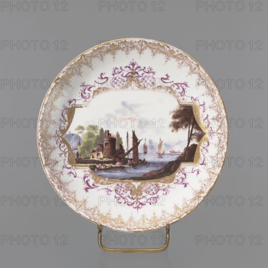 Jug plate, Koenigliche Porzellain Fabrique, Meissen circa 1720–1735, Meissen, Saxony. Porcelain shallow bowl plate in an oval, concave-convex, multi-part reserve in a mirror, bordered with gilded lace and supplemented with purple leafy volutes with fields of Chinese lattice. Central colorful landscape miniature with staffage: a bay with a sailing boat, architecture including a round building on the shore, and two sailing boats in the distance. Around the rim a trail of gilded, serrated lace. On the reverse of the collar three Indianische Blumen branches. Height 3.8 cm; diameter 18 cm. Maker attribution: Koenigliche Porzellain Fabrique, Meissen 1710–1763., A round ceramic plate with a central painted scene framed by ornate decorative borders; the scene shows a waterfront with sailing boats and a small stone building near trees, and a shoreline with a tree on one side, painted in muted tones of brown, green, blue, and gray with touches of reddish-brown; the plate rim is decorated with in...
