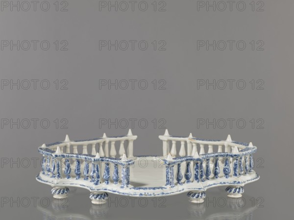 Base for a tabletop tray. Oval architectural tray serving as a base for a tabletop tray. Carved with 4 leaves, with a wavy edge — alternately concave and convex. Supported by 8 round, spirally fluted legs. Around the edge is a baluster gallery with balusters interwoven with small columns and pillars, topped with cones. Painted in a blue Regency arabesque. Inner walls and base white. 1st half of the 19th century; possibly 18th century France, Strasbourg . Vessel. Height 11.0 cm; width 33.0 cm; depth 45.0 cm., A decorative ceramic object with a pale white base and blue painted ornamentation sits on a reflective surface; it has a low, irregularly shaped platform surrounded by a balustrade of small turned posts and pointed finials, with the blue decoration forming floral and linear patterns along the rim, posts, and rounded feet, and the overall color scheme includes white and blue against a gray background.