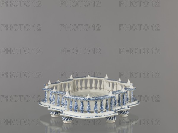 Base for a tabletop tray. Oval architectural tray base carved with 4 leaves and a wavy edge alternately concave and convex. Supported by 8 round, spirally fluted legs. Around the edge is a baluster gallery with balusters interwoven with small columns and pillars, topped with cones. Painted in a blue Regency arabesque; inner walls and base white. 1st half of the 19th century; possibly 18th century France, Strasbourg. Vessel. Height 11.0 cm, width 33.0 cm, depth 45.0 cm., A small decorative ceramic stand with a round base supported by a group of short turned feet, painted in white with blue painted floral and scroll motifs, featuring a raised balustrade-like rim with several turned spindles and pointed finials, all reflected faintly on a glossy gray surface with a uniform gray background.