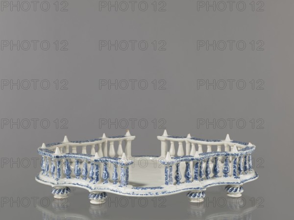 Base for a tabletop tray. Oval architectural tray serving as a base for a tabletop tray. Carved with 4 leaves, with a wavy edge—alternately concave and convex. Supported by 8 round, spirally fluted legs. Around the edge is a baluster gallery with balusters interwoven with small columns and pillars, topped with cones. Painted in a blue Regency arabesque. Inner walls and base white. 1st half of the 19th century; 18th century, France, possibly Strasbourg. Vessel. Height 11.0 cm, width 33.0 cm, depth 45.0 cm., A shallow decorative dish with an open lattice-like rim sits on a reflective surface; it is white with blue painted floral and scroll motifs, has a scalloped edge, pointed finials along the rim, turned baluster-shaped supports around the perimeter, and rests on small twisted column feet, all visible against a plain gray background.