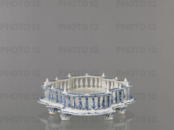 Base for a tabletop tray. Oval architectural tray serving as a base for a tabletop tray. Carved with 4 leaves and a wavy edge—alternately concave and convex. Supported by 8 round, spirally fluted legs. Around the edge is a baluster gallery with balusters interwoven with small columns and pillars, topped with cones. Painted in a blue Regency arabesque. Inner walls and base white. 1st half of the 19th century; 18th century, France, Strasbourg . Vessel. Height 11.0 cm; width 33.0 cm; depth 45.0 cm., A ceramic dish with a decorative openwork rim resembling a small fence sits on a reflective surface, showing a glossy white base color with detailed blue painted floral and scroll motifs on the rim, baluster-shaped supports, and rounded feet; the rim has pointed finials and the interior is smooth and white, while the background is a uniform gray.