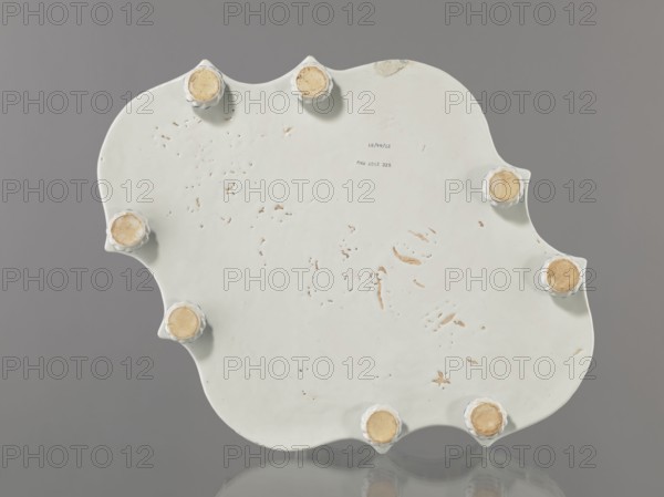 Base for a tabletop tray. Oval architectural tray serving as a base for a tabletop tray. Carved with 4 leaves, with a wavy edge—alternately concave and convex. Supported by 8 round, spirally fluted legs. Around the edge is a baluster gallery with balusters interwoven with small columns and pillars, topped with cones. Painted in a blue Regency arabesque. Inner walls and base white. 1st half of the 19th century; 18th century, France, Strasbourg ; vessel. Height 11.0 cm, width 33.0 cm, depth 45.0 cm., A pale off-white scalloped ceramic object viewed from the underside resting on a reflective surface, with a smooth slightly worn surface showing small chips and scratches and a few scattered brownish spots; several raised rounded feet with beige worn pads are arranged around the edge; the background and reflection are shades of gray.