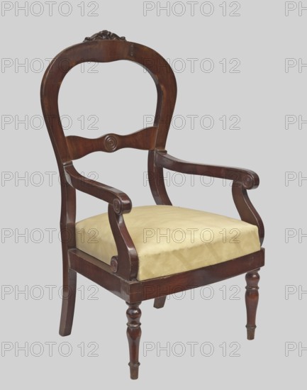 Armchair, veneered with mahogany, with a trapezoidal upholstered seat removable cushion and a balloon-shaped openwork backrest fastened in the middle with a decorative crossbar with a wavy outline. The front legs are turned and profiled; the rear legs are angular and arched backwards. The armrests are S-shaped with volute ends, supported by S-shaped supports. The finial features carved acanthus leaves; the centre of the backrest crossbar and the volutes of the armrests and their supports feature decorative, profiled knobs. Styles: Louis Philippe, Biedermeier. Materials/ornament: plant decoration, knobs. Date: 1835–1845. Place of origin: Warsaw . Function: seating furniture. Dimensions: height 114.8 cm, width 63 cm, depth 59.5 cm., A wooden armchair with a curved, carved backrest and scrolled armrests, featuring turned front legs and straight back legs, a flat wooden frame, and a cushioned seat covered in a light beige fabric; visible colors include dark brown wood tones, light beige up...