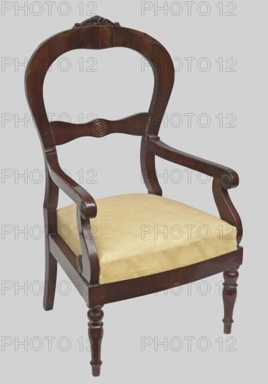 Armchair, plant decoration, knobs ornament, Biedermeier style, Louis Philippe style. ARMCHAIR veneered with mahogany, with a trapezoidal upholstered seat removable cushion and a balloon-shaped openwork backrest fastened in the middle with a decorative crossbar with a wavy outline. The front legs are turned, profiled; the rear legs are angular, arched backwards. The armrests are S-shaped with volute ends, supported by S-shaped supports. The finial features carved acanthus leaves; the centre of the backrest crossbar and the volutes of the armrests and their supports feature decorative, profiled knobs. 1835–1845. Possibly Warsaw. Piece of furniture, seating piece. Height 115.5 cm, width 63 cm, depth 59 cm., A wooden armchair with a curved backrest and carved crest, a carved horizontal back slat with a round carved detail, scrolled armrests with carved circular accents, a padded seat upholstered in a pale yellow fabric, turned front legs and straight back legs, all set against a light gray...