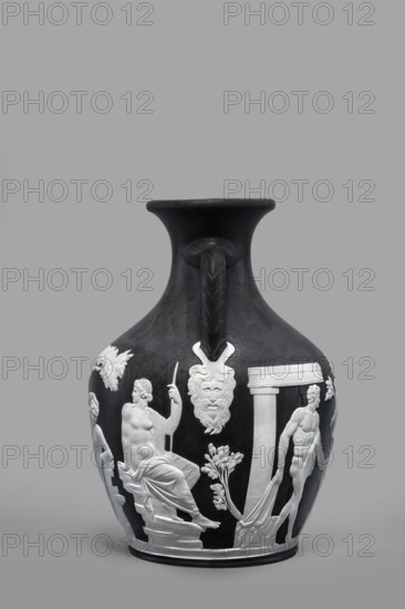 Pelike-shaped vase, copy of the Portland Vase by Josiah Wedgwood & Sons Stoke-on-Trent pottery manufactory, 1790–1895. Black body with white cameo-style reliefs rendered from a black background depicting: 1 Atia mother of Emperor Augustus, Apollo, and Neptune; 2 Hecuba wife of Priam holding a torch, surrounded by Venus and Paris; and at the bottom, Paris lost in thought. 19th/20th century, Staffordshire, Stoke-on-Trent, England. Height 26.6 cm; width 19 cm. Maker: Wedgwood, Josiah 1730–1795., A dark-colored ceramic jug with a wide flared neck and a single curved handle is shown against a neutral gray background; the body of the jug features raised white relief figures and decorative elements, including a seated draped male figure holding a staff, a standing partially draped male figure, a theatrical mask with a beard, architectural column-like shapes, floral motifs, and flowing drapery, with visible colors being black, white, and shades of gray.