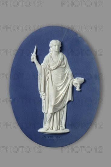 Plaque with Melpomene, Etruria Factory; Flaxman the Younger, John after; Wedgwood, Josiah; Lewinski, Jan Piotr — mythologies; jasper stoneware; oval sapphire plaque with white relief overlay depicting the muse of tragedy, Melpomene, with the inscription MELPOMONE embossed below; relief after the design of John Flaxman showing Apollo and nine Muses; rectangular mahogany wood frame; ca. 1780–1785; England, Staffordshire, Stoke-on-Trent., A white sculpted relief of a robed female figure is set against a blue oval background, the figure holding a small knife in one hand and a shallow bowl on a stem in the other, wearing draped clothing with visible folds and sandals, with pale skin tones in the sculpture and surrounding colors of blue and gray.