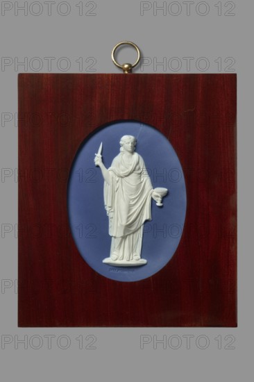 Plaque with Melpomene, Etruria Factory Staffordshire; pottery factory; 1769–1950. Flaxman the Younger, John after, Wedgwood, Josiah 1730–1795 factory owner, Flaxman, John 1755–1826, Wedgwood, Josiah 1730–1795. Mythologies, jasper stoneware. Oval sapphire plaque with white relief overlay — muse of tragedy, Melpomene, with the inscription MELPOMONE embossed below; relief after the design of John Flaxman: Apollo and nine Muses. In a rectangular mahogany wooden frame. ca. 1780–1785. England; Staffordshire; Stoke-on-Trent. Plaque., A rectangular wooden plaque in a deep reddish-brown tone with a brass hanging ring at the top surrounds an oval indented panel in a muted blue color, within which a white relief sculpture of a robed standing figure is visible holding a short blade in one hand and a shallow cup in the other, the figure showing draped clothing and detailed folds.