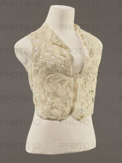 Bodice, lace, provenance, bodices; women's clothing, fashion. In a pattern of rosettes connected by large spiders and heads; pattern of stylized floral motifs. 19th century, France. Clothing accessory; height 36 cm, circumference 63 cm., A dress form mannequin in an off-white color displays a sleeveless lace vest in a pale cream shade; the vest features intricate openwork floral and circular lace patterns and scalloped edges, with a low V-shaped front and a small fabric tie at the center, and the background is a muted grayish-brown.