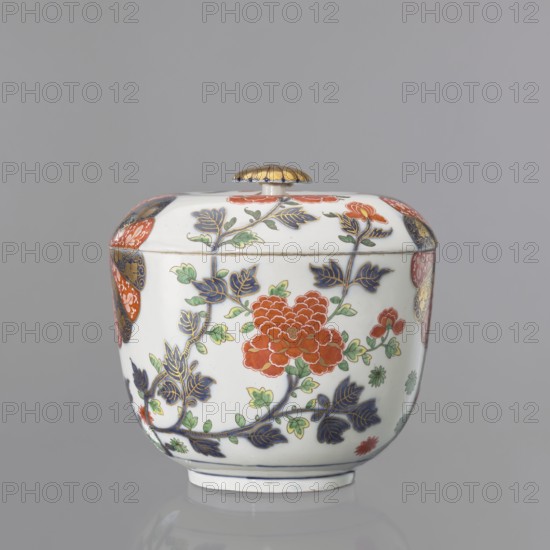 Vase with lid with Imari decoration, Kaiserliche Porzellanmanufaktur in Wien 1744–1864. Hemispherical vase with a separated bottom rim. Flat-arched lid with a button-rosette handle. Around the body, in a quadripartite arrangement, is Imari decoration continuing onto the lid: two fan-shaped 14-petal rosettes in brick red and cobalt blue with gilding, alternating with a fan-shaped cluster of lanceolate leaves with lotus flowers at the base of the bottom rim and two smaller tufts of leaves on the sides, and a peony shoot-branch with two full brick-red flowers and two buds; between the four motifs at the bottom are green and brick-red star-flowers and small leaves. Gilded rosette handle with a cobalt disc and a brick-red volute around the base. At the bottom, a double cobalt border with an inscribed signature in the center. Mid-19th century. 18th century, Austria, Vienna. Decorative vessel; height 20.5 cm, diameter 21.5 cm., A lidded ceramic vessel sits against a neutral gray background, s...