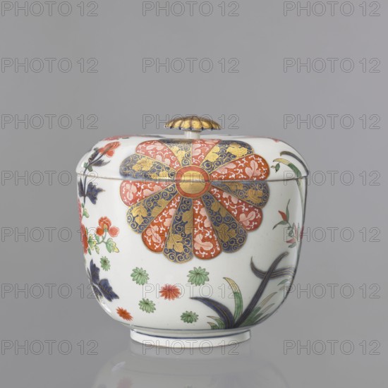 Vase with lid with Imari decoration, Kaiserliche Porzellanmanufaktur in Wien 1744–1864. Maker attributed to Praglowski, Jan fl. ca. 1954. Hemispherical vase with a separated bottom rim and a flat-arched lid with a button-rosette handle. Around the body, in a quadripartite arrangement, is Imari decoration continuing onto the lid: two fan-shaped 14-petal rosettes in brick red and cobalt blue with gilding, alternating with a fan-shaped cluster of lanceolate leaves with lotus flowers at the base of the bottom rim and two smaller tufts of leaves on the sides, and a peony shoot-branch with two full brick-red flowers and two buds; between the four motifs at the bottom are green and brick-red star-flowers and small leaves. Gilded rosette handle with a cobalt disc and a brick-red volute around the base. At the bottom, a double cobalt border with an inscribed signature in the center. Mid-19th century. 18th century, Austria; Vienna. Decorative vessel. Height 20.5 cm, diameter 21.5 cm., A lidded c...