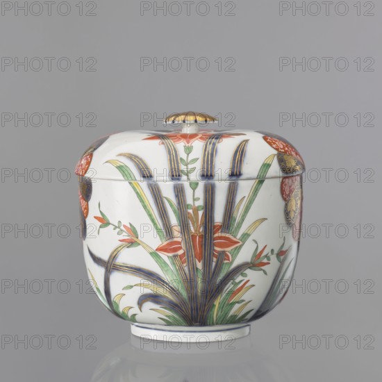Vase lid with Imari decoration, Kaiserliche Porzellanmanufaktur in Wien 1744–1864; Imari style, Far Eastern-style floral decorations. Flat-arched lid with Imari decoration continued on the vase in a 4-part arrangement: two 6-petal brick-red cobalt half-rosettes with gilding, a lotus flower with leaves, and a peony flower on a branch with a small bud. Flat gilded button-rosette handle set on the lid in a cobalt band with a brick-red spiral. Mid-19th century; 18th century; Vienna, Austria. Decorative vessel; height 5 cm, diameter 21.5 cm., A lidded ceramic jar with a rounded body and a small knob handle on top sits against a plain gray background; the jar is predominantly white and decorated with a floral motif featuring a group of tall, slender leaves in green and blue with gold accents, several orange-red flower shapes, and additional green foliage, while the lid edge and sides show a few circular decorative panels in red, blue, and gold; the knob on the lid is gold-toned and the base ...