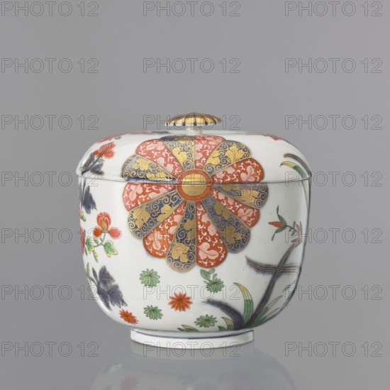 Vase lid with Imari decoration, Kaiserliche Porzellanmanufaktur in Wien 1744–1864. Imari style, Far Eastern–style floral decorations. Flat-arched lid with Imari decoration continued on the vase in a 4-part arrangement: two 6-petal brick-red cobalt half-rosettes with gilding, a lotus flower with leaves, and a peony flower on a branch with a small bud. Flat gilded button-rosette handle, set on the lid in a cobalt band with a brick-red spiral. Mid-19th century–18th century, Vienna, Austria. Decorative vessel. Height 5 cm; diameter 21.5 cm. Maker attribution: Praglowski, Jan fl. ca. 1954 removed as collection provenance., A rounded lidded porcelain container sits against a plain gray background, predominantly white with painted floral and foliate decoration. The lid has a small domed handle with a gold and darker detailing. A large rosette motif adorns the front, composed of alternating petal shapes in red and blue with gold ornamentation radiating from a central circular element. Smaller ...